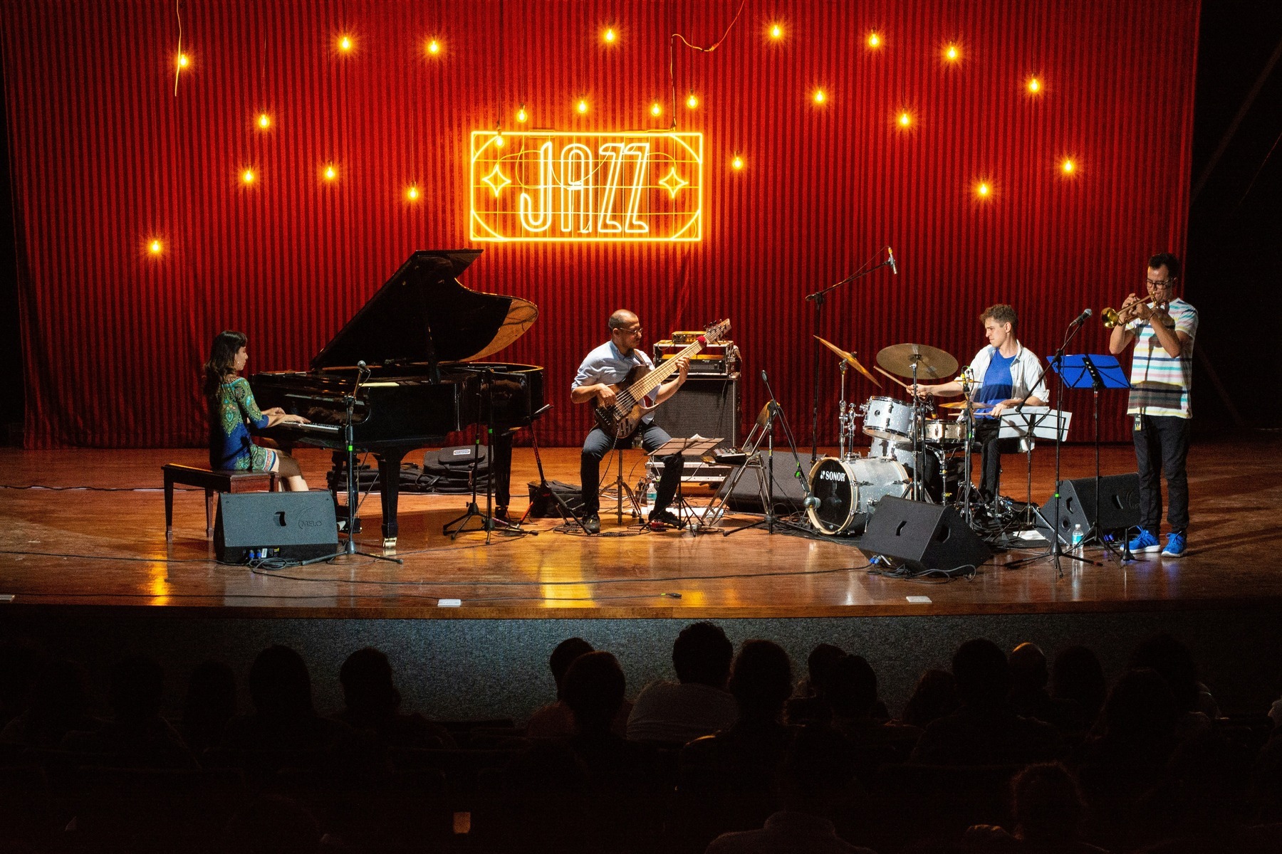 A jazz band performing on stage beneath a glowing 'JAZZ' sign as a seated audience watches