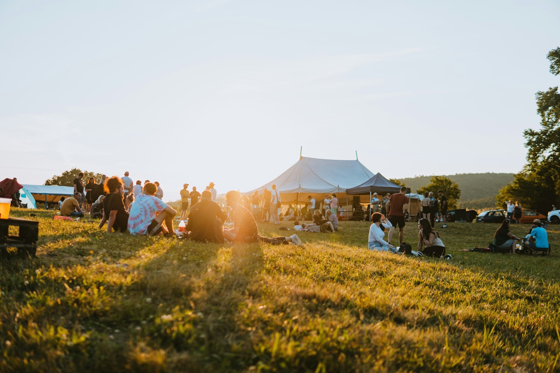 Festival-goers relaxing on the grass at golden hour beside an open-air event tent