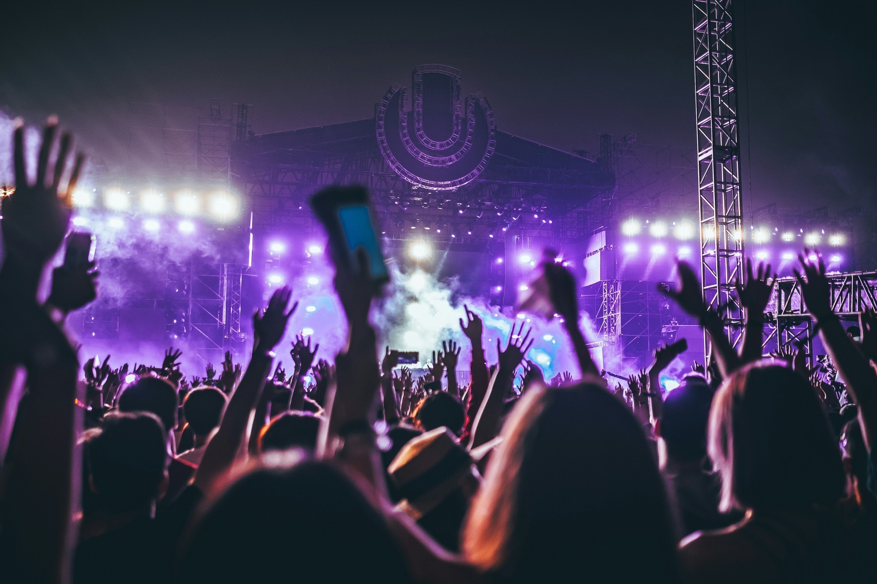 A festival crowd with raised hands in front of a brightly lit main stage at night