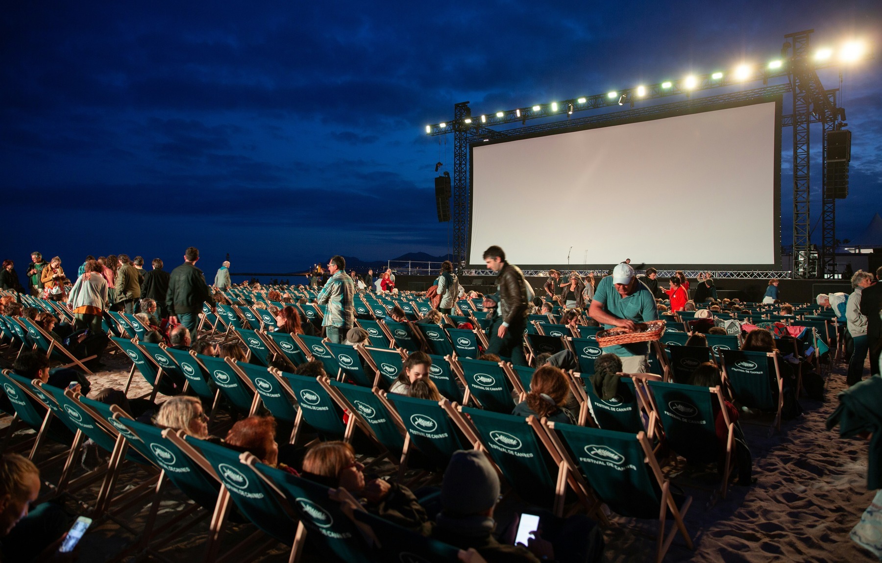 A large open-air cinema screen glowing at dusk above rows of deckchairs and an evening audience