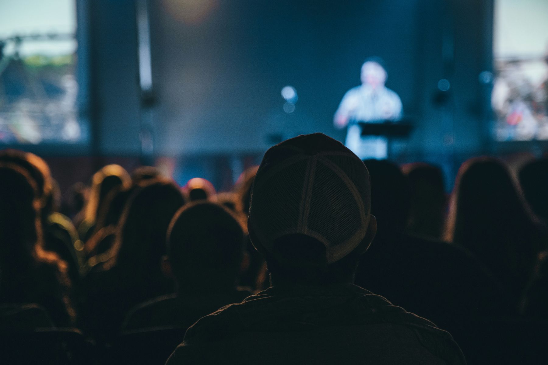 An attendee in a cap watching a keynote speaker on stage at a conference