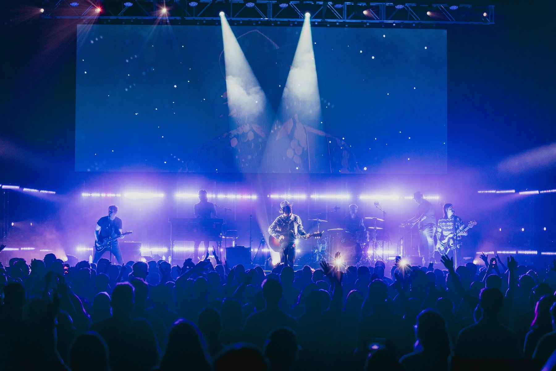 A rock band performing under blue stage lights to a packed indoor arena crowd