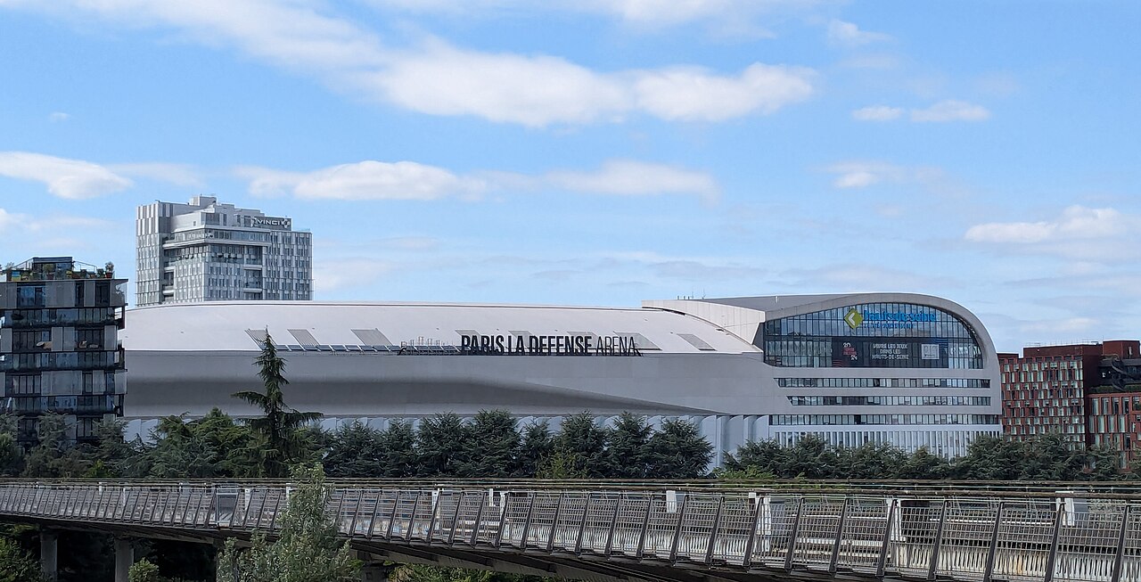 The Paris La Défense Arena seen by day, with its curved white facade rising above the footbridge in Nanterre